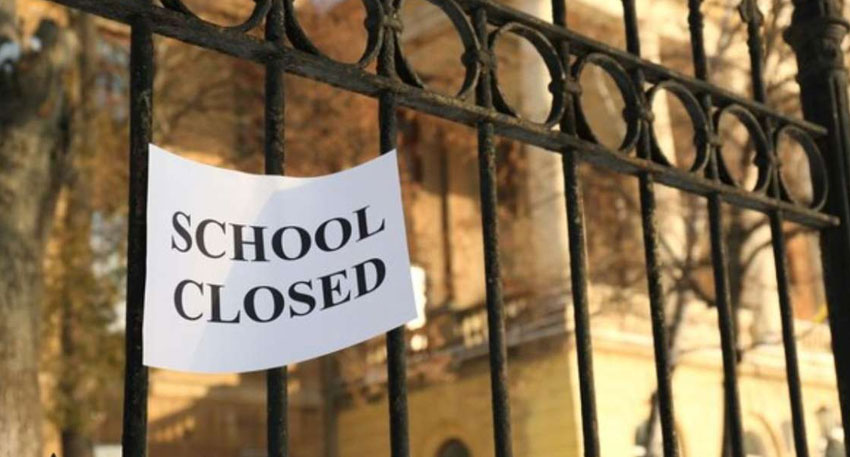 A paper sign reading “School Closed” hangs on a black iron gate outside a school building
