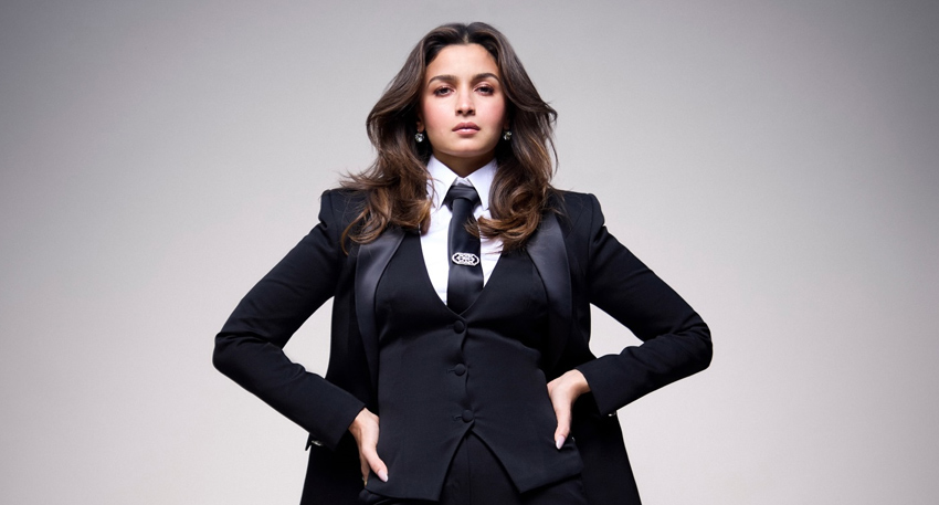 Alia Bhatt in a black three-piece suit with a tie and white shirt, posing with hands on her hips.