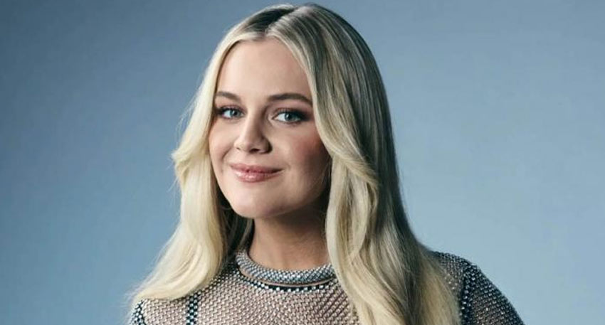 Kelsea Ballerini smiling in a mesh top with blonde hair against a blue background.