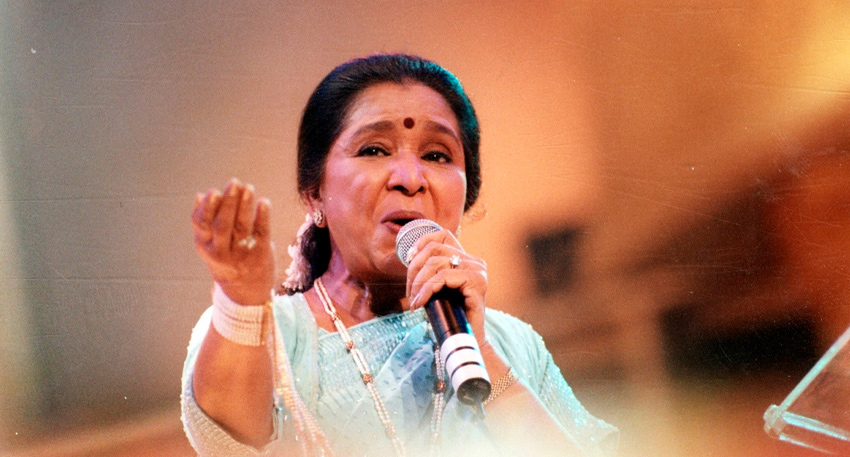 Legendary Indian playback singer Asha Bhosle performing live on stage with a microphone.