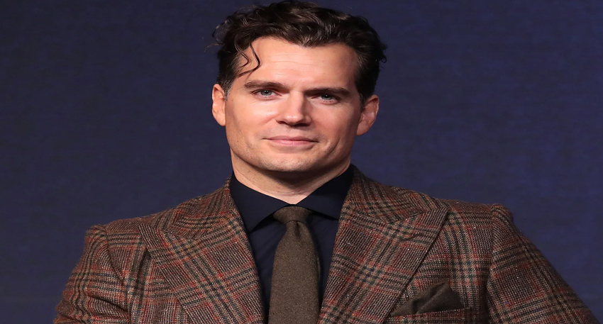 Henry Cavill wearing a patterned brown suit and tie against a dark blue background.