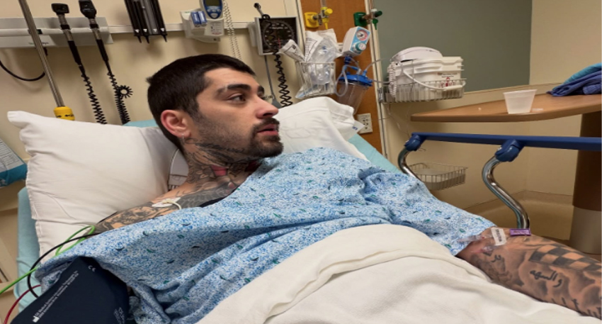 Zayn Malik in a hospital bed wearing a gown, looking to the side with medical monitors and an IV visible.