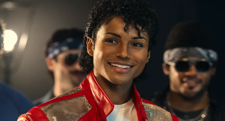 Jaafar Jackson as Michael Jackson, wearing a red and gold military jacket, smiling warmly in a dark room.