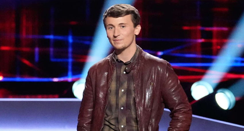 Singer Dylan Carter smiling in a brown leather jacket on 'The Voice' stage with red and blue lights behind him.