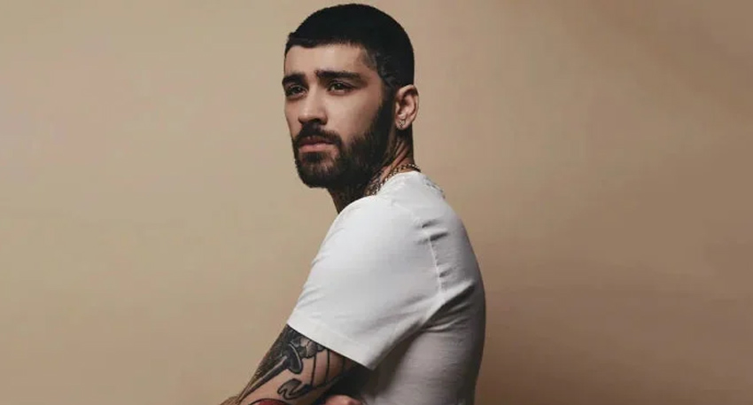 Profile shot of Zayn Malik with a buzz cut and beard, wearing a white t-shirt against a tan background.