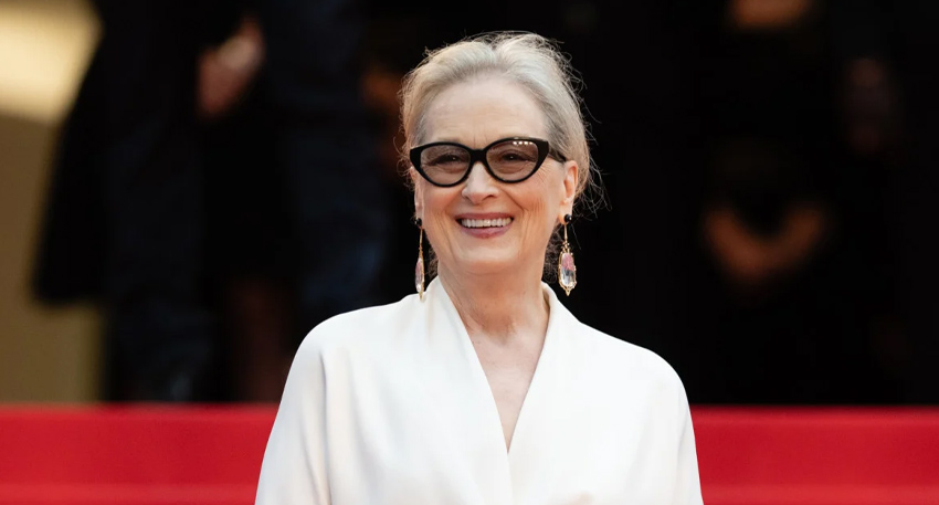 Meryl Streep smiling on a red carpet wearing a white dress, black-rimmed glasses, and long drop earrings.
