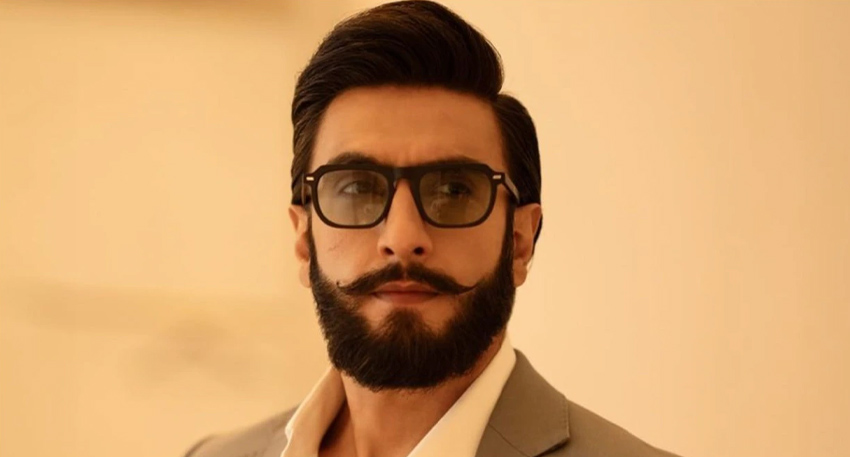 Bollywood actor Ranveer Singh wearing thick black glasses and a tailored grey suit jacket.