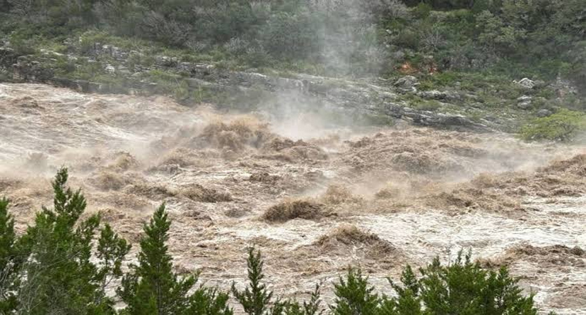 Raging brown floodwaters surge violently through a rocky river valley lined with green trees and mist.