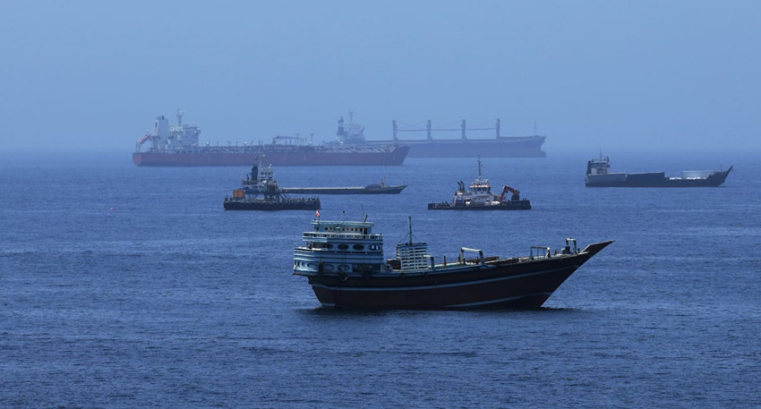 : Commercial cargo vessels and a traditional dhow boat anchored in the Strait of Hormuz waterway.