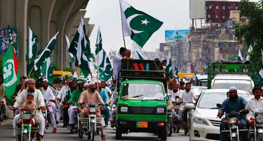 Crowded street rally in Pakistan with people on motorcycles and a truck carrying large Pakistani flags.