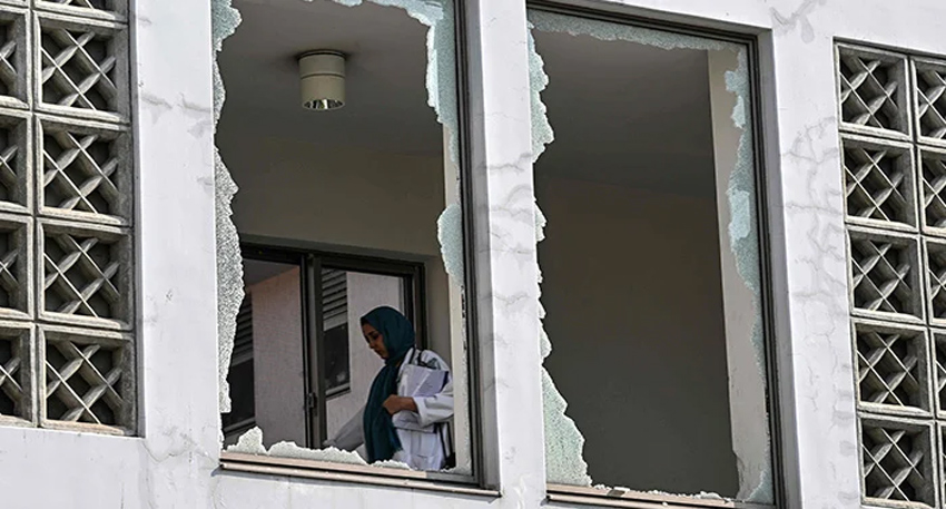 A hospital worker stands inside a building behind a shattered glass window showing fire damage.