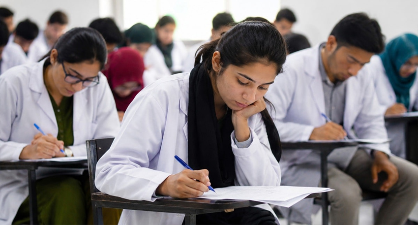 Medical students in lab coats sitting at desks and taking an exam in a classroom.