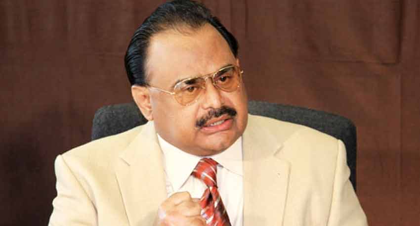 Pakistani politician Altaf Hussain wearing a cream-colored suit, a striped red and white tie, and gold-rimmed glasses, sitting and clenching his fist.