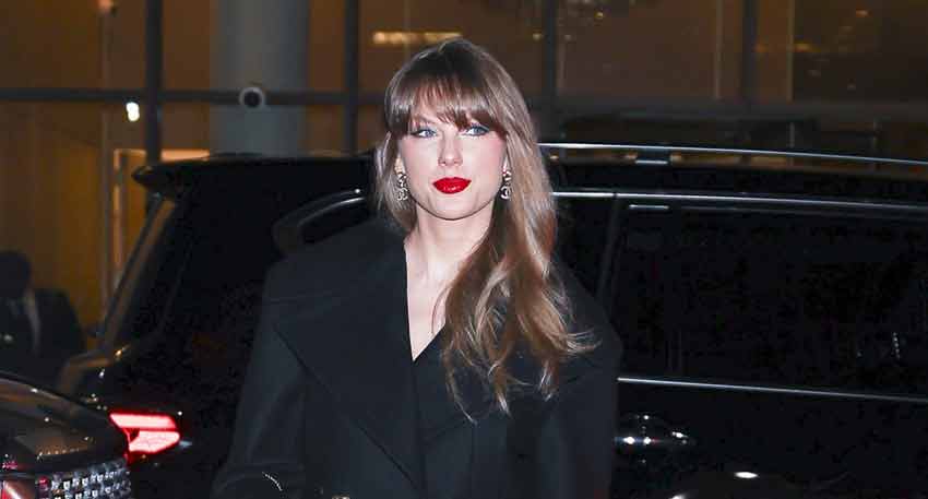 Taylor Swift wearing a black coat and red lipstick, standing outdoors next to a black SUV at night.