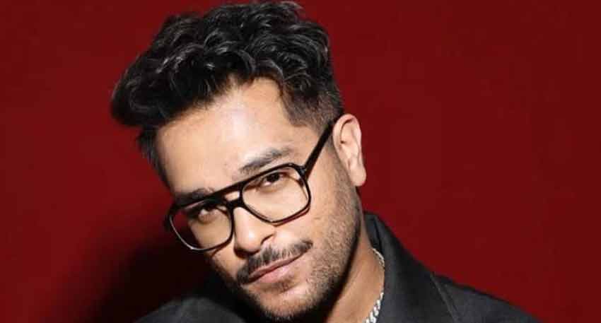 Asim Azhar, a Pakistani singer and actor, poses for a photo wearing black-rimmed glasses against a red background.