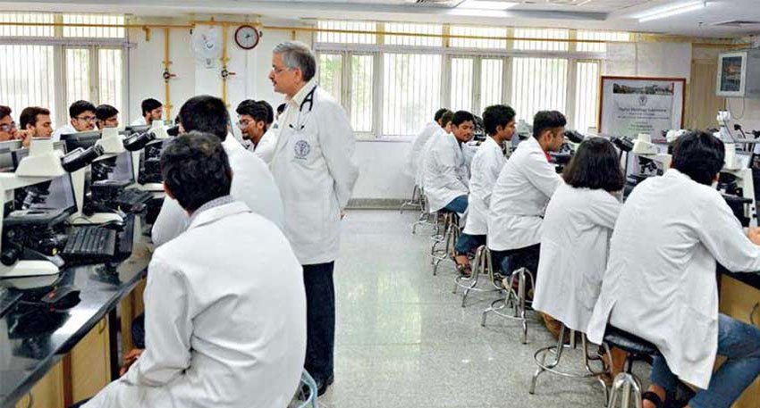 Medical students in white lab coats work with microscopes in a busy classroom as an instructor observes.