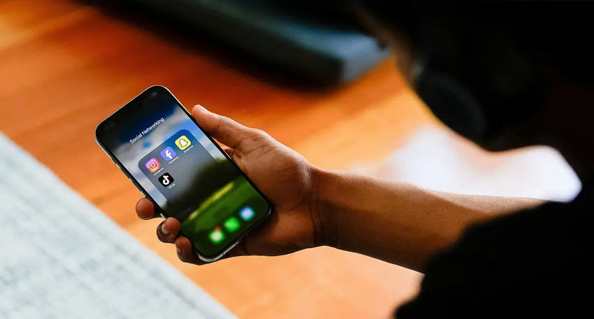 A person holds a smartphone displaying a folder of social networking apps including Instagram, Snapchat, Facebook, and TikTok.