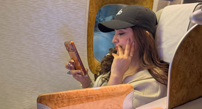 Hina Niazi, wearing a black cap, sits in an airplane seat next to a window, looking at her phone. Photo: Hina Niazi Facebook