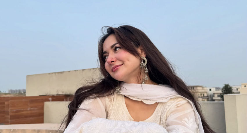 Hania Aamir wearing a white outfit and silver earrings, smiles while looking away from the camera on a rooftop. Photo: Hania Aamir Instagram