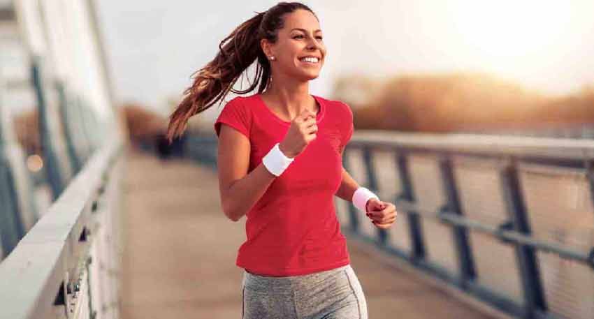 Smiling woman with a ponytail in a red shirt and grey shorts jogging on a bridge at sunset