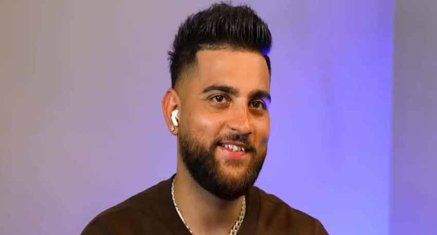Karan Aujla, a Punjabi singer, smiling and wearing earbuds and a silver chain, in a studio setting.