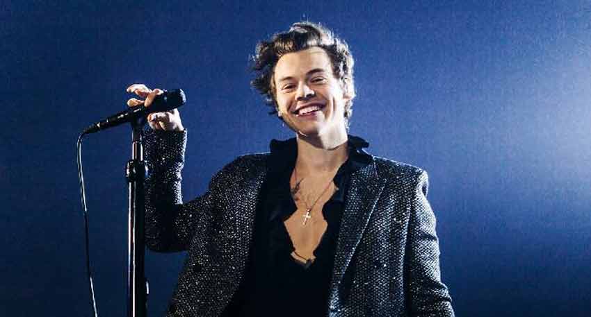 Harry Styles on stage, smiling and holding a microphone, wearing a sparkly jacket and a black ruffled shirt.