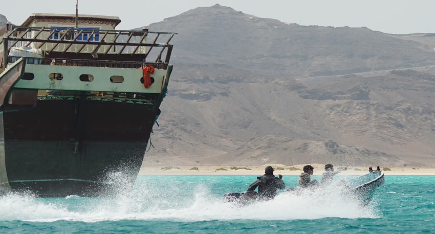 A small skiff speeds past a large vessel near a rocky coastline.