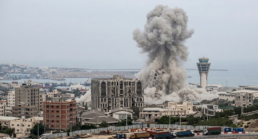 A large explosion cloud rises from a building in a coastal city near a control tower and port area.