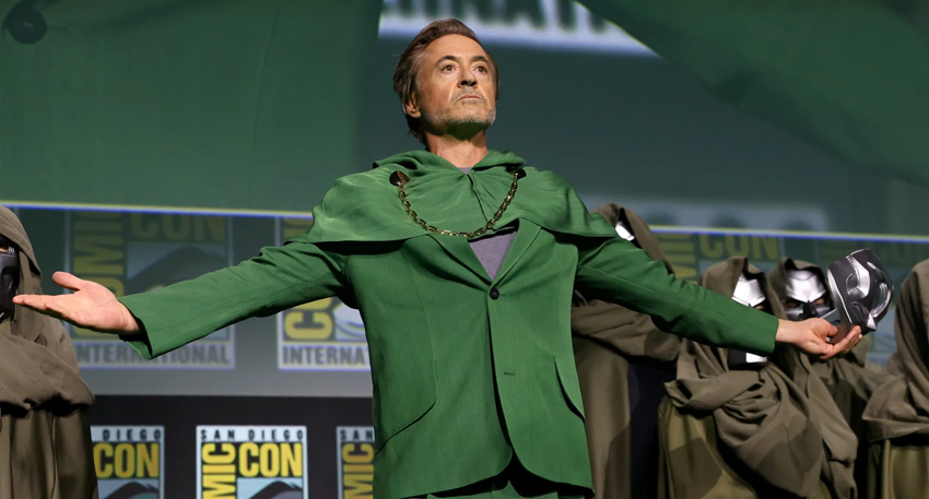 Robert Downey Jr. wearing a green suit and cape on stage at Comic-Con holding a Doctor Doom mask.