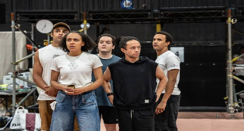 Five actors standing together on a rehearsal stage looking upward with expressions of shock and worry.