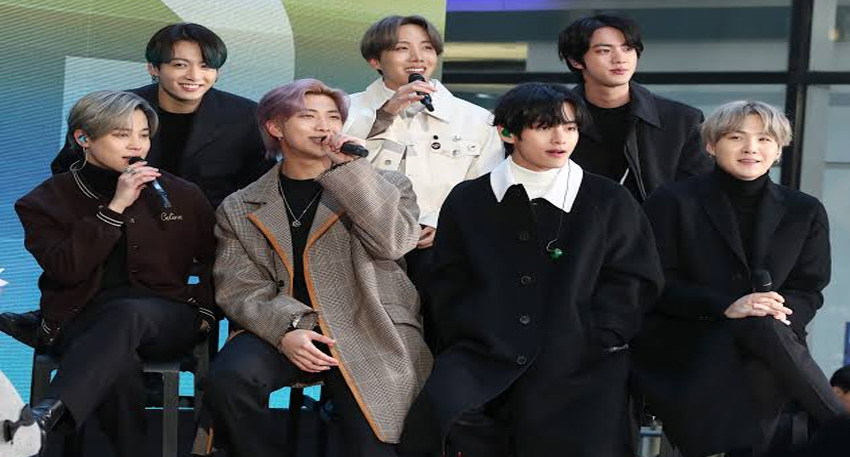 Seven members of BTS sitting together and smiling during a live television performance.