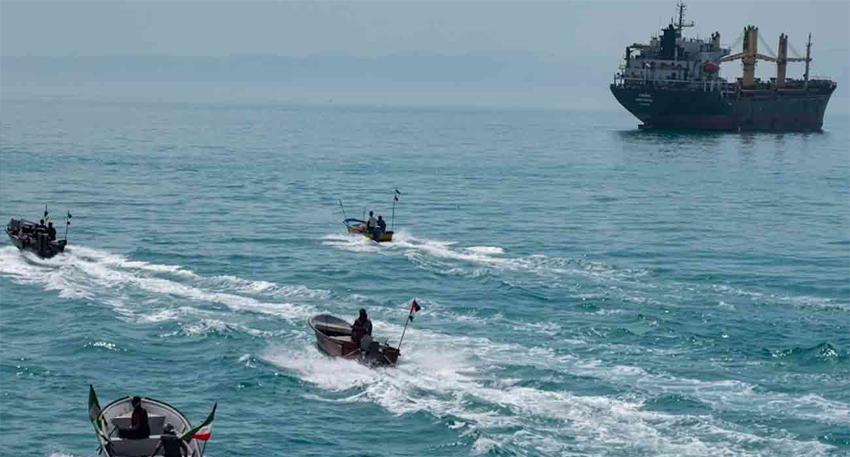 Four Iranian speedboats surrounding a large cargo ship in the turquoise waters of the Strait of Hormuz.
