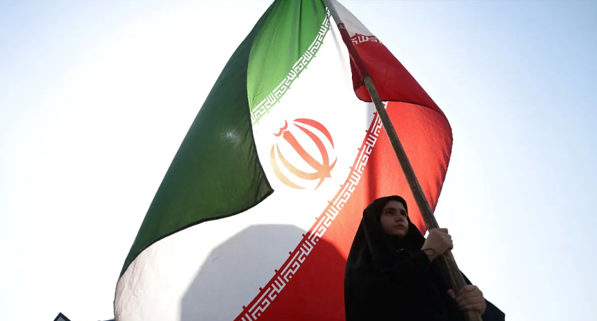 Iranian woman in black holding a large Iranian flag against a clear, bright sky.