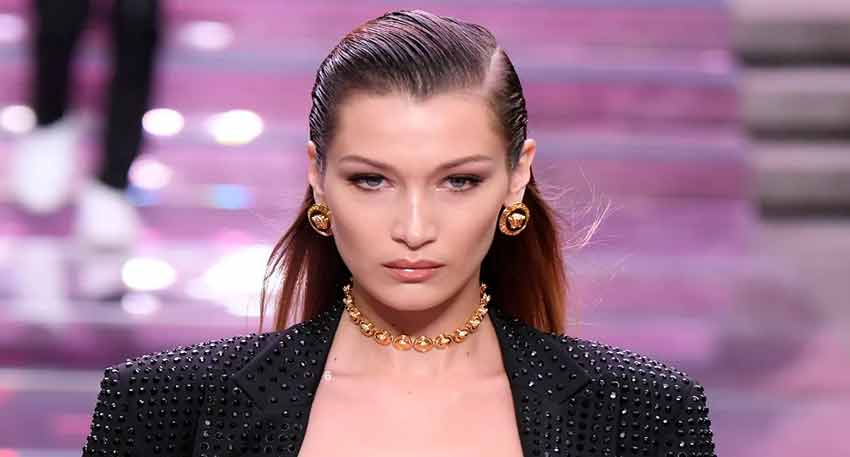 Model Bella Hadid on the Versace runway wearing a black studded blazer and gold jewelry with wet-look hair.