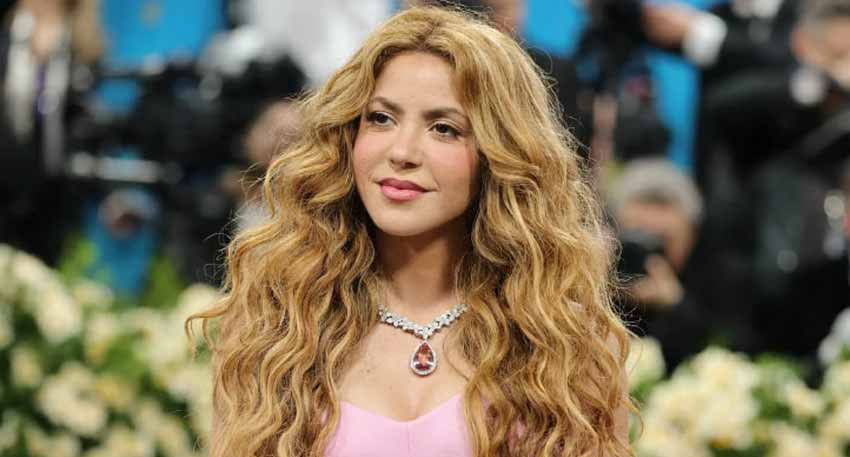 Shakira with long, wavy blonde hair wearing a pink dress and a large diamond and ruby necklace.