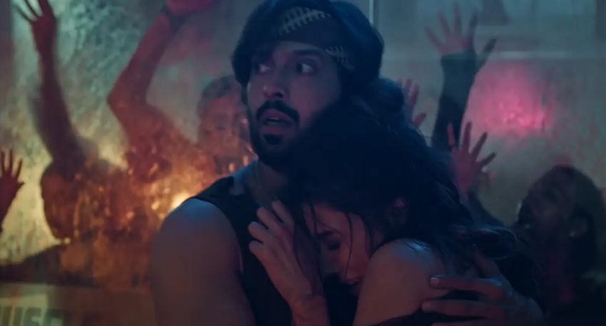Fahad Mustafa in a headband and black tank top holds a woman, Mehwish Hayat, tightly as zombies swarm a glass window behind them.