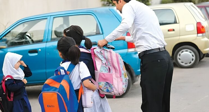 Schoolchildren wearing heavy backpacks while a man helps adjust one beside a blue car in a parking lot.