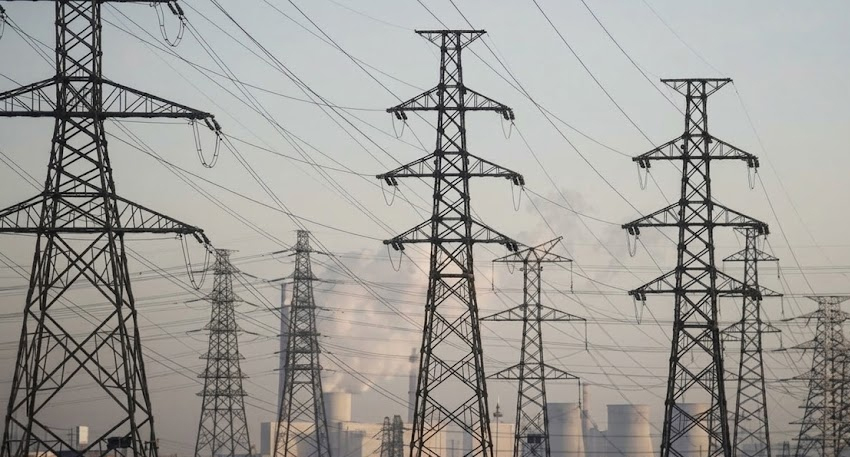Lattice transmission towers and power lines against a hazy sky with industrial cooling towers in the background.