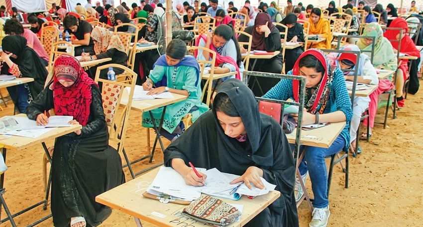 Pakistani students seated at wooden desks in an outdoor examination hall, writing their tests.