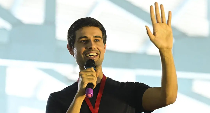 Indian YouTuber Dhruv Rathee holding a microphone and waving his hand while speaking on stage.