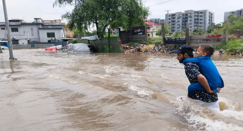 A man carrying a child on his back walks through deep floodwater on an urban street after heavy rain.