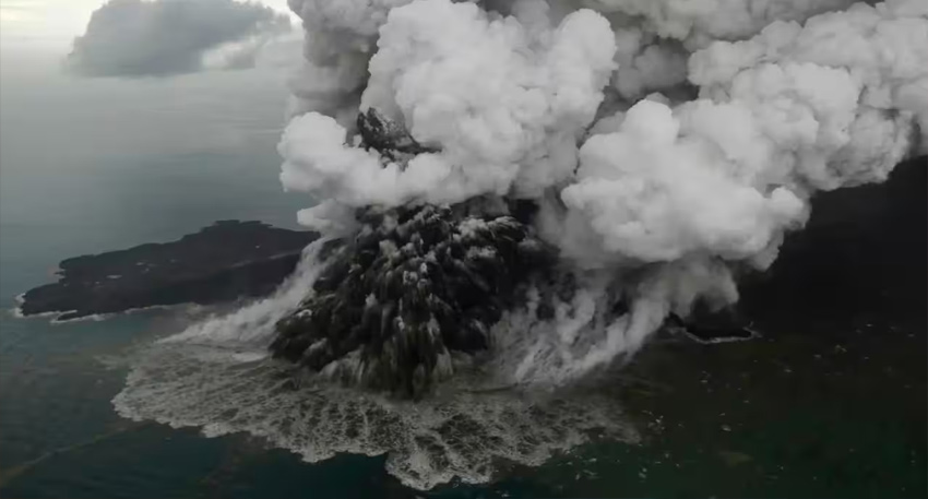 An aerial view of an island volcano violently erupting, sending dense white ash clouds into the sky and water.