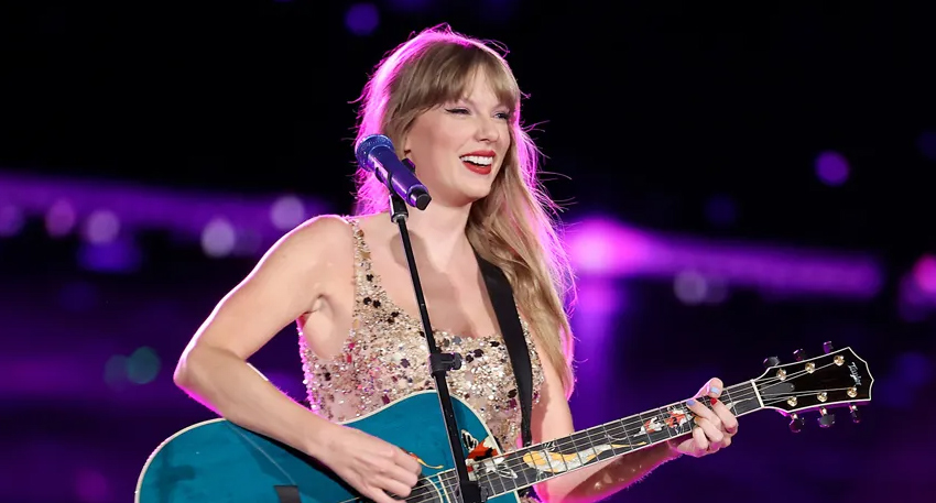 Taylor Swift smiling and playing a teal acoustic guitar on stage during the Eras Tour.
