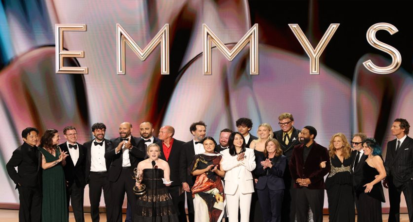 The cast and crew of a winning show celebrate on stage under a large 