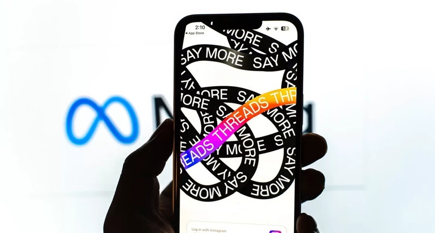 Hand holding a smartphone displaying the Threads app logo with a Meta logo blurred in the background.