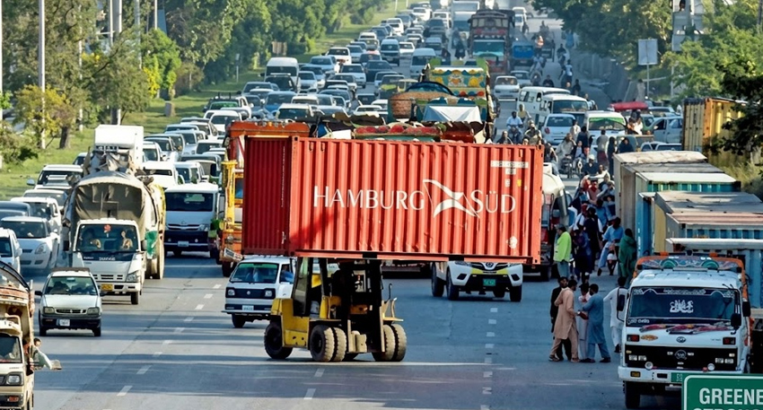 A forklift carrying a large red shipping container blocks a heavily congested highway filled with cars and trucks.