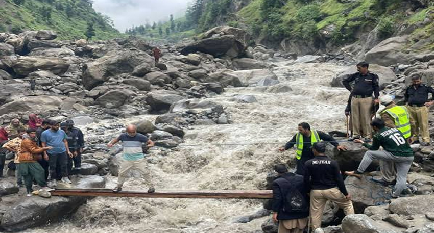 Rescue workers help people cross a raging, muddy river over a makeshift wooden plank bridge in a rocky valley.